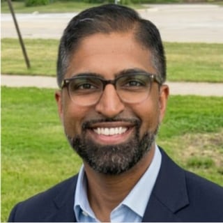 Portrait of a smiling man with dark beard and glasses wearing a navy blazer over a light blue shirt, outdoors with green grass background